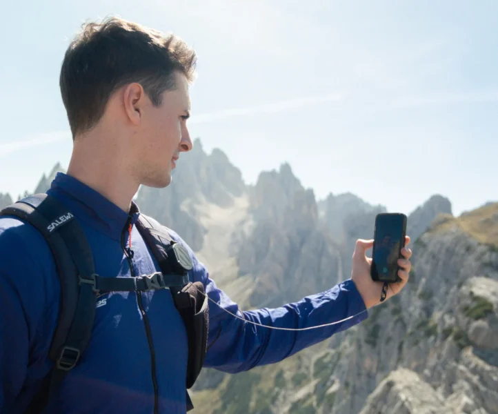 Ein junger Mann hält sein durch ein Seil gesichertes Smartphone im ausgestreckten Arm vor einer Bergkulisse.
